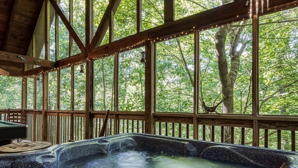A hot tub area with a wooden deck and large windows overlooking a forest. - Mountain Setting Large Screen in Back Porch offers Pool table and Hot Tub Private Setting (Sevierville)