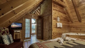 A cozy log cabin bedroom with a large bed, a TV, a wooden ceiling, and a view of the outdoors. - Mountain Setting Large Screen in Back Porch offers Pool table and Hot Tub Private Setting (Sevierville)