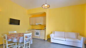 Una habitación luminosa de color amarillo con una mesa de comedor, sillas, un sofá y una zona de cocina con armarios y una estufa.