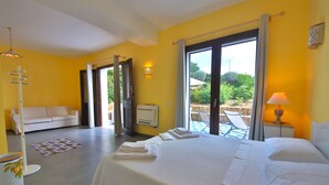 Habitación de hotel con cama, sofá, mesita de noche con lámpara, ventana con cortinas y vistas al exterior.