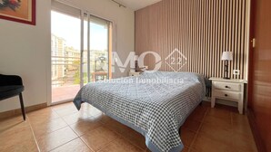 A bedroom with a large bed, a nightstand, a lamp, a chair, and a view of buildings through a sliding door. - 1st line beach & terrace with sea views (Cambrils)