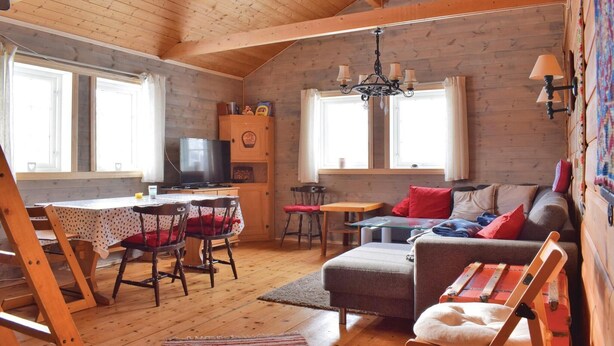 Ein gemütliches Blockhauszimmer mit Holzbalkendecke, einem Fernseher, einer Couch, einem Esstisch mit Stühlen und einer kleinen Küchenzeile.