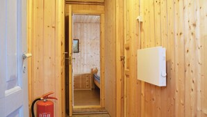 Ein schmaler Holzkorridor mit einem Feuerlöscher links, einem Bett mit Streifenmuster im Hintergrund und einem Thermostat an der rechten Wand.