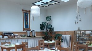 Een eetgedeelte met houten tafels en stoelen, een grote plant en een keuken op de achtergrond.