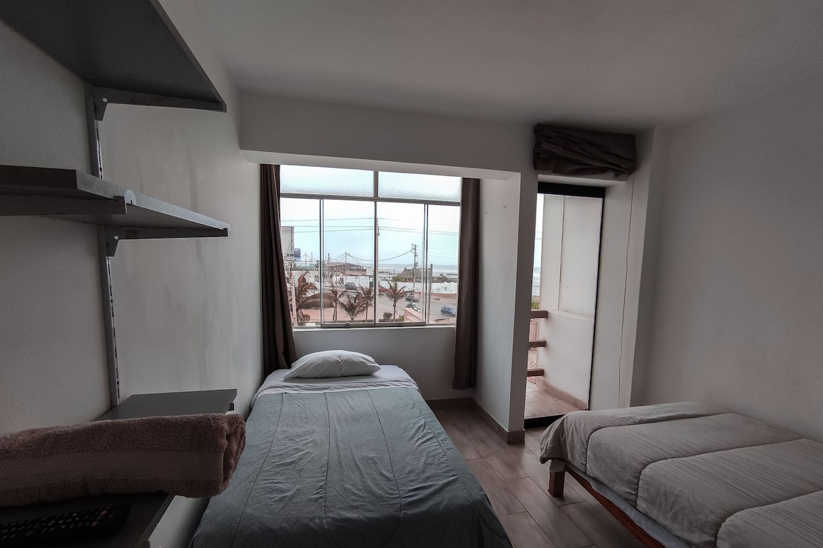 A hotel room with a bed, a desk with a keyboard, and a window with a view of palm trees and buildings.