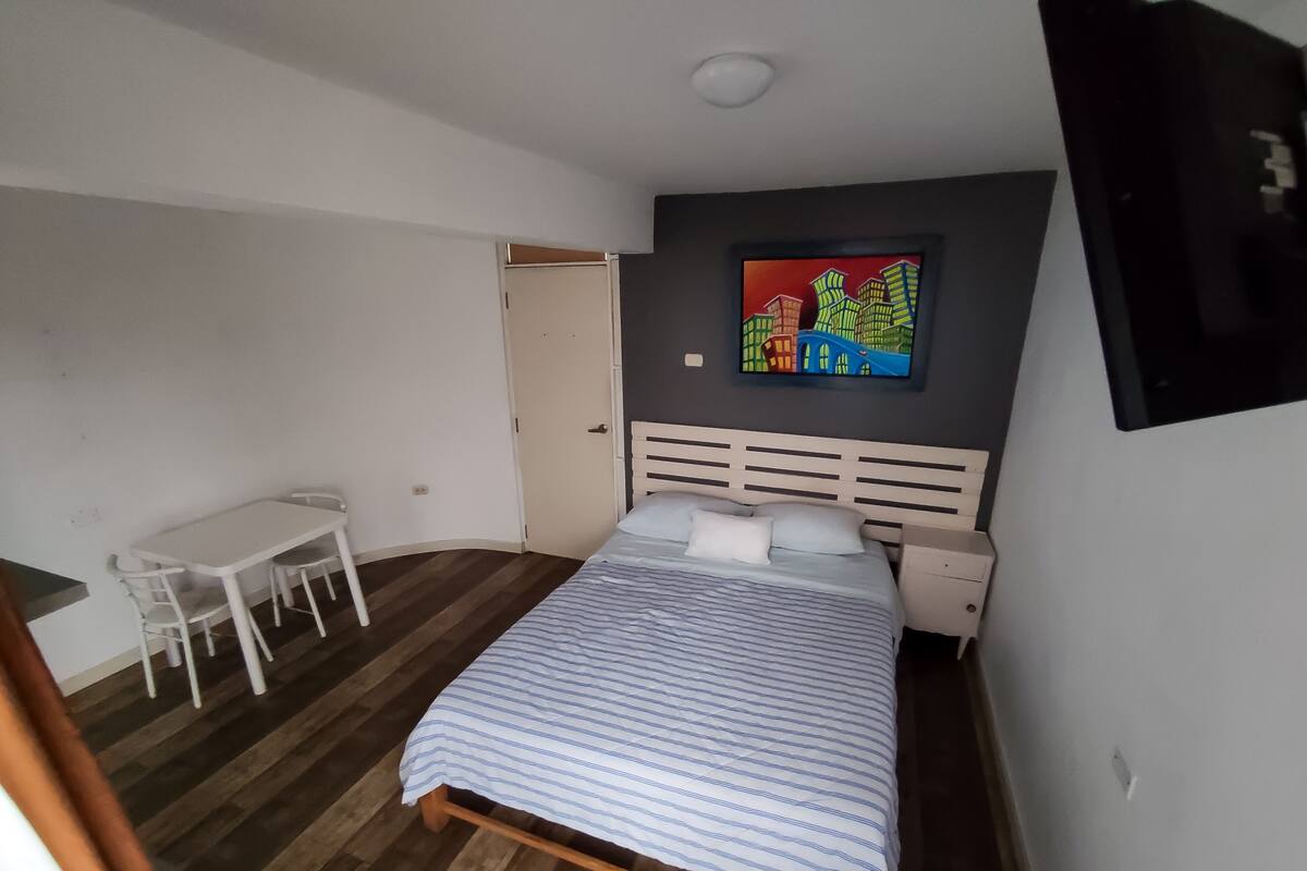 A bedroom with a bed, a desk, and a chair. There is a painting on the wall and a door leading to another room.