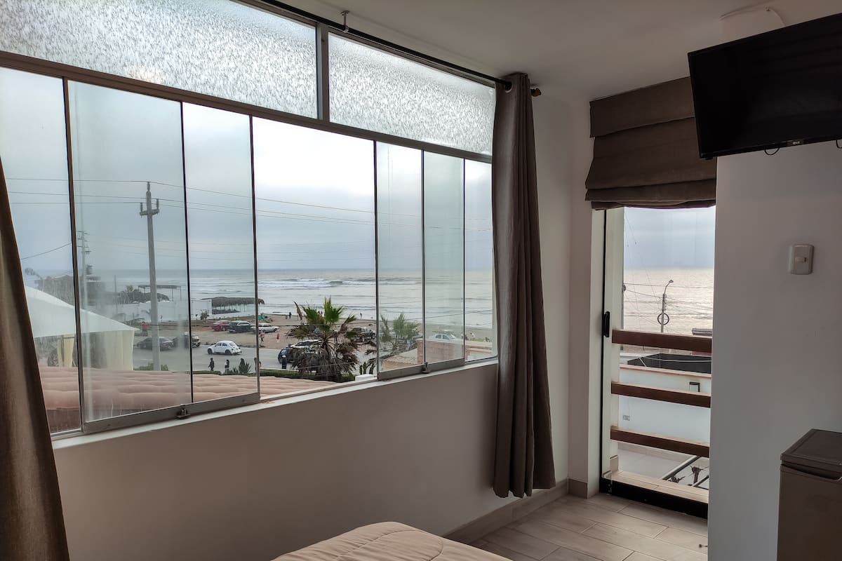 A room with a large window overlooking a beach, a bed, and a TV.
