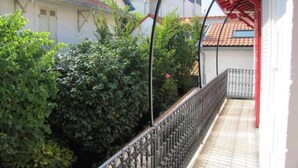 Un balcón techado con baranda de metal, rodeado de abundante vegetación y edificios vecinos con tejados rojos.