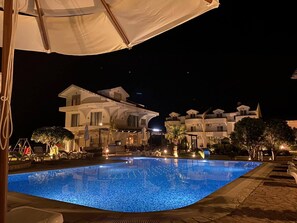 Une piscine éclairée pour la baignade nocturne, avec un grand bâtiment en arrière-plan, entourée d’espaces de détente en plein air et de palmiers.