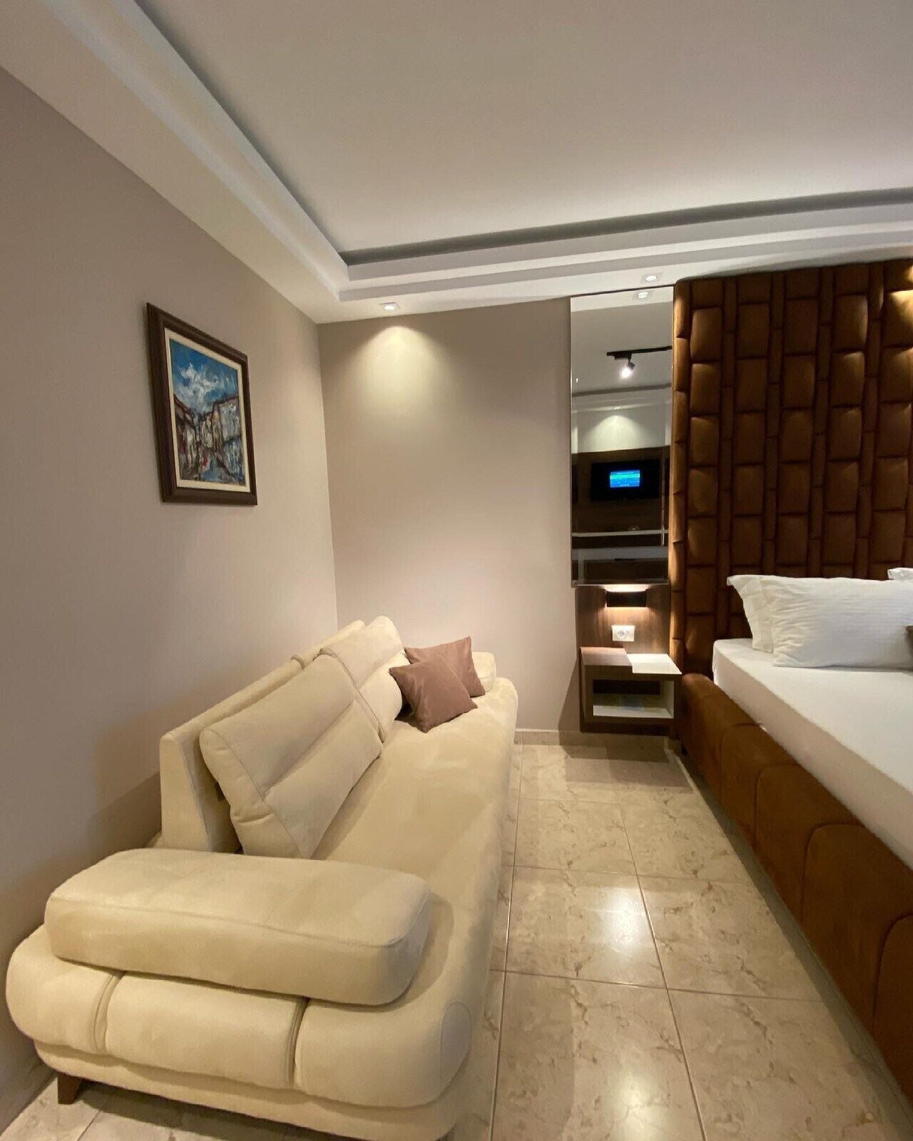 A modern hotel room with a beige sofa, a bed with a headboard, a flat-screen TV, and a small table.