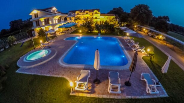 Una gran piscina con sillas de descanso y sombrillas, rodeada por un césped bien iluminado y un edificio al fondo.