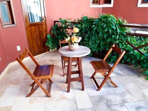 Un patio con una mesa redonda y dos sillas de madera, rodeado de macetas con plantas y un muro rojo.