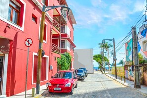 Un edificio rojo con un letrero de ‘no estacionar’, un coche rojo estacionado al frente y una calle adoquinada.