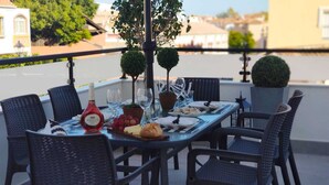 A table set for dining on a balcony with wicker chairs, a bottle of Mateus wine, and a view of the cityscape.