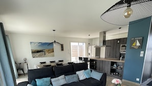 Un salon moderne avec un canapé d’angle foncé, une table basse en bois ornée de fleurs et un grand luminaire suspendu noir.