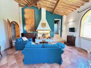 A living room with a fireplace, blue sofas, a wooden ceiling, and a large tapestry. - Luxury apartment in Tuscan villa with pool: your own window to iconic Siena! (Siena)