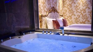 Een groot, rechthoekig bubbelbad met meerdere kraanwerk, voor een muur met een spiegel die een bed met een gestoffeerd hoofdeinde en behang met patroon weerspiegelt.