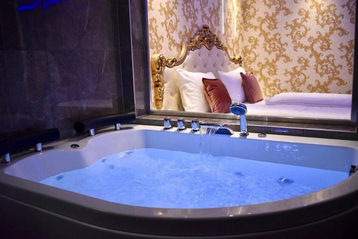 Un grand bain à remous rectangulaire, doté de plusieurs robinets, installé contre un mur orné d’un miroir qui reflète un lit avec une tête de lit capitonnée et un papier peint à motifs.