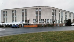 Een groot, rechthoekig gebouw met een plat dak en meerdere ramen. Er is een overdekte ingang met een bord erboven.