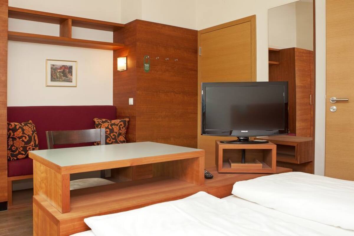 A hotel room with a wooden bed, a TV on a wooden stand, a wooden desk, and a red cushioned bench.