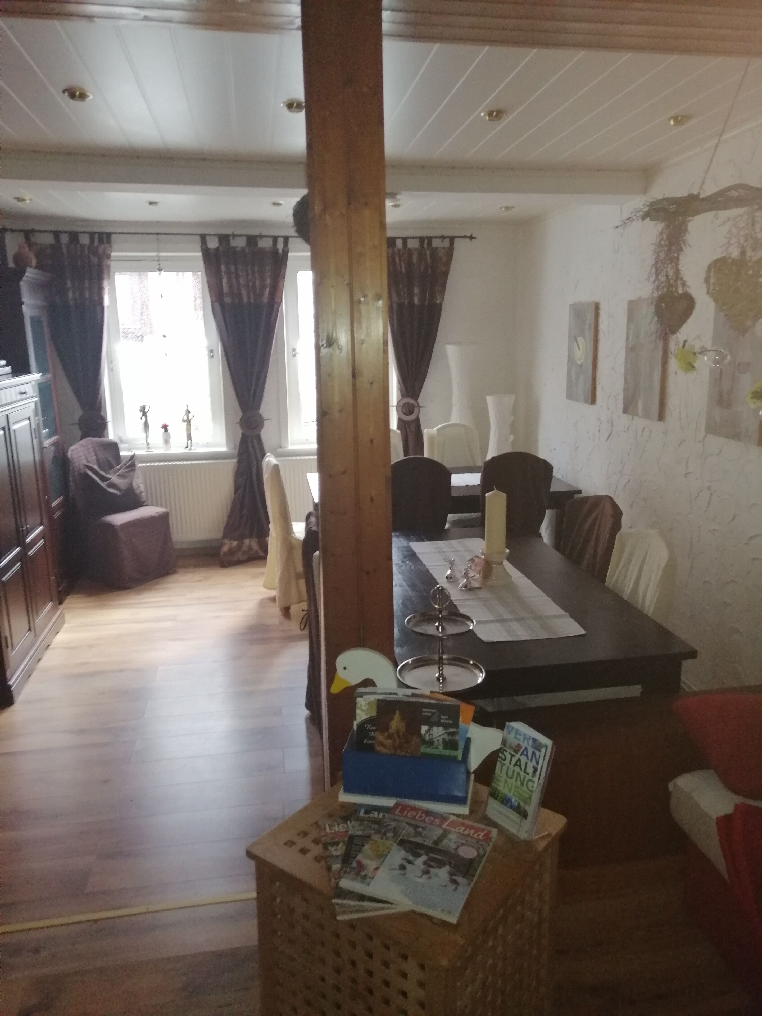 Ein Essbereich mit einem Holztisch, Stühlen und einem Fenster mit Vorhängen. Eine Korbwicker-Kiste mit Zeitschriften und ein kleiner Tisch mit einer Kerze stehen dort.