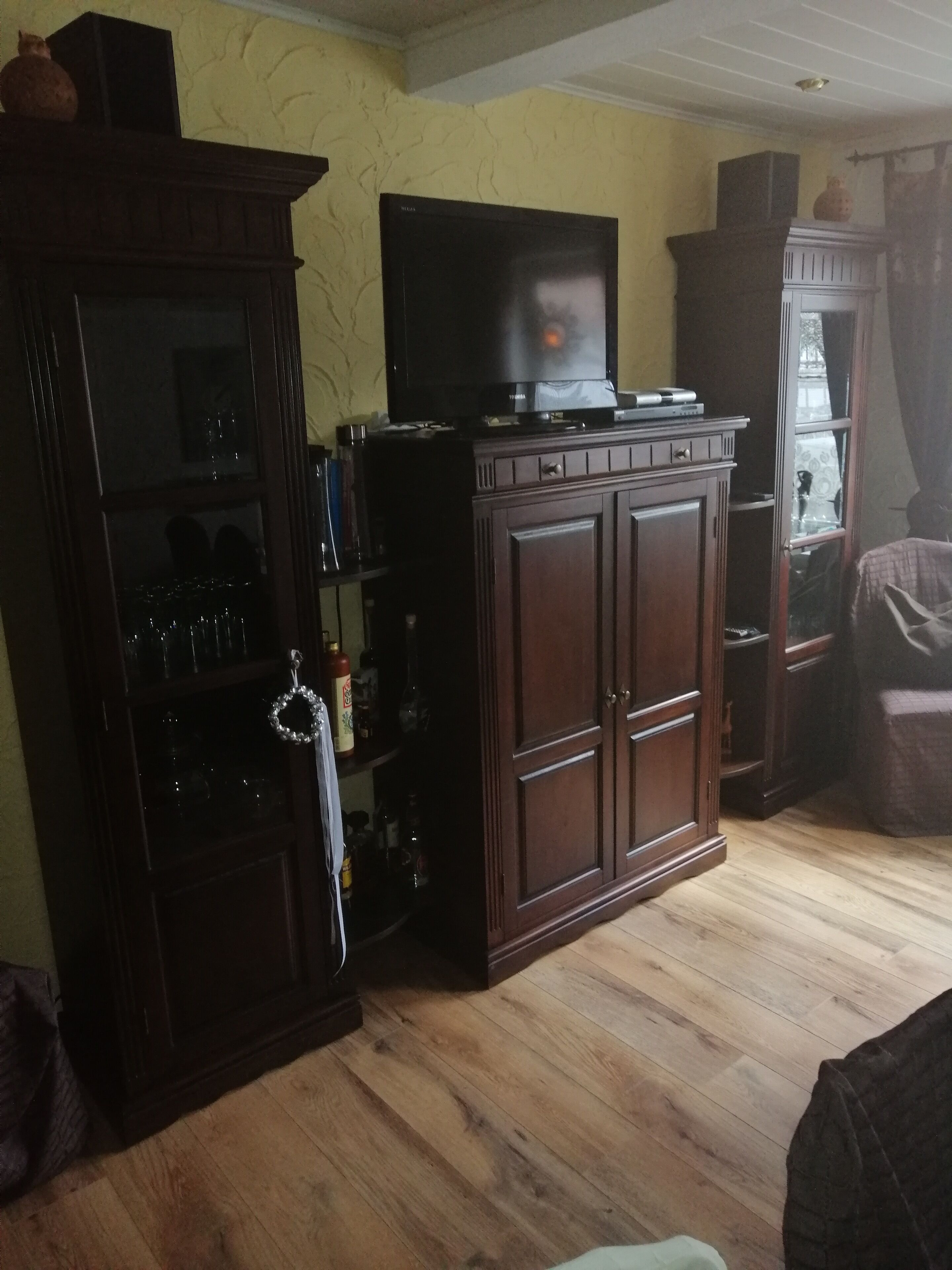 A living room with wooden cabinets, a television on top, and a glass cabinet with bottles.