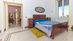 Un dormitorio con cama de madera, colcha con estampado azul y blanco, mesita de noche de madera, una mesilla auxiliar, una ventana con vistas y una puerta que da a otra habitación.