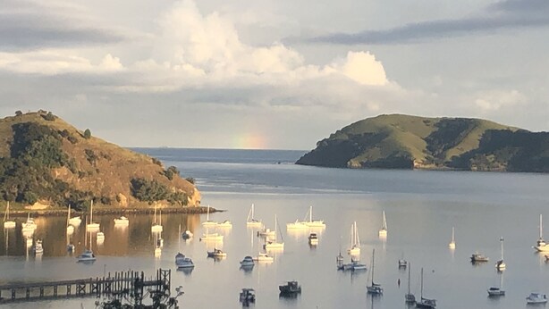 Cena costeira com barcos atracados num cais, um arco-íris ao longe e colinas ao fundo.