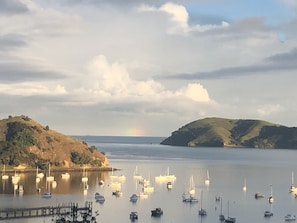 Cena costeira com barcos atracados num cais, um arco-íris ao longe e colinas ao fundo.