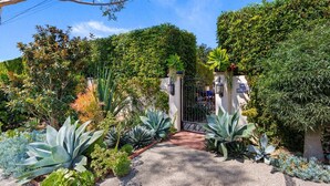 A gated entrance with a paved path leading to a building, surrounded by various plants and trees.