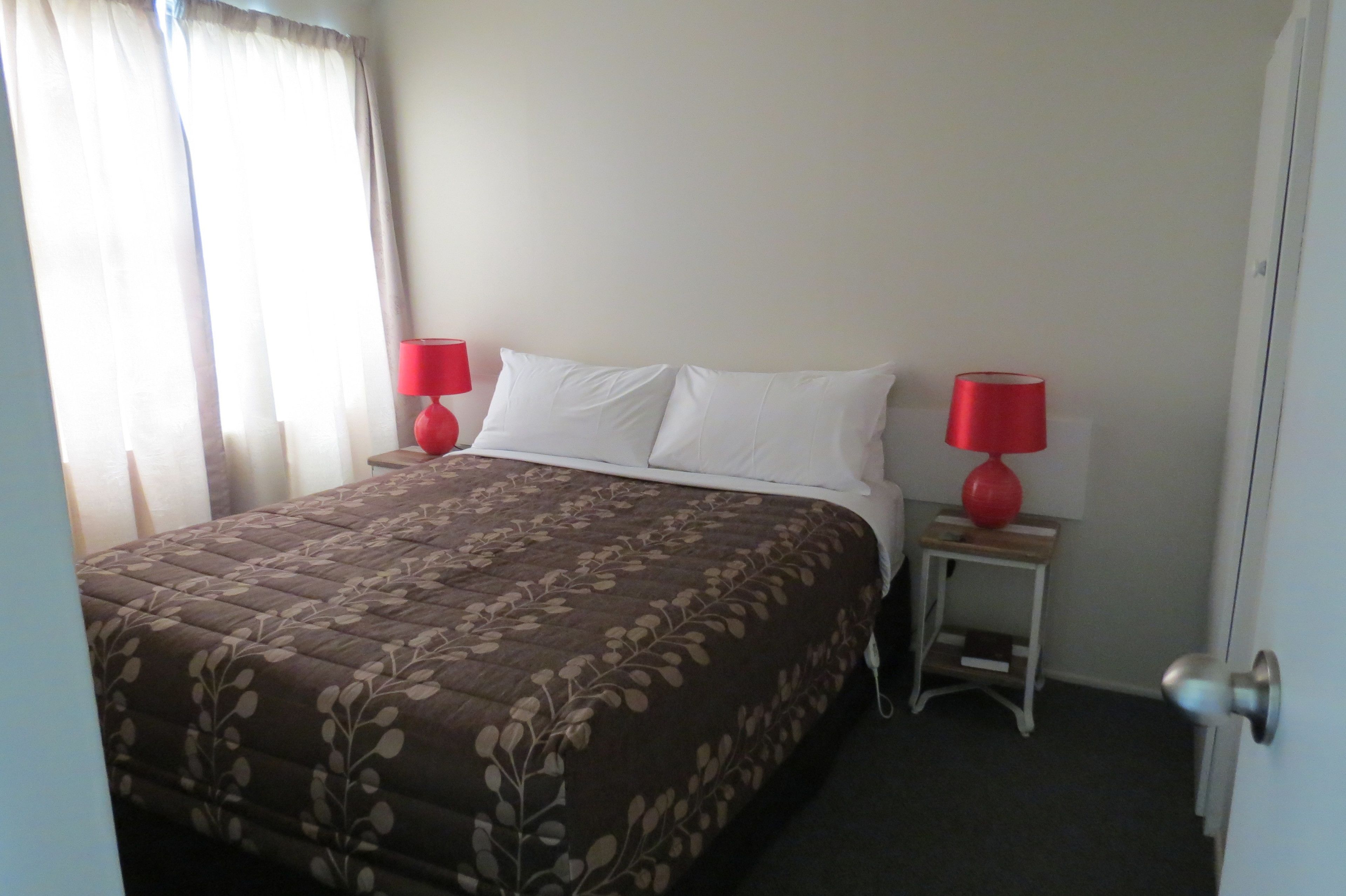 Una cama bien hecha con una colcha floral marrón, dos lámparas de noche rojas y una mesita de noche de madera con un jarrón rojo.
