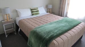 Una cama bien hecha con una manta verde, almohadas blancas y una cubierta acolchada color beige. Hay dos mesitas de noche con lámparas y un pequeño taburete de madera.