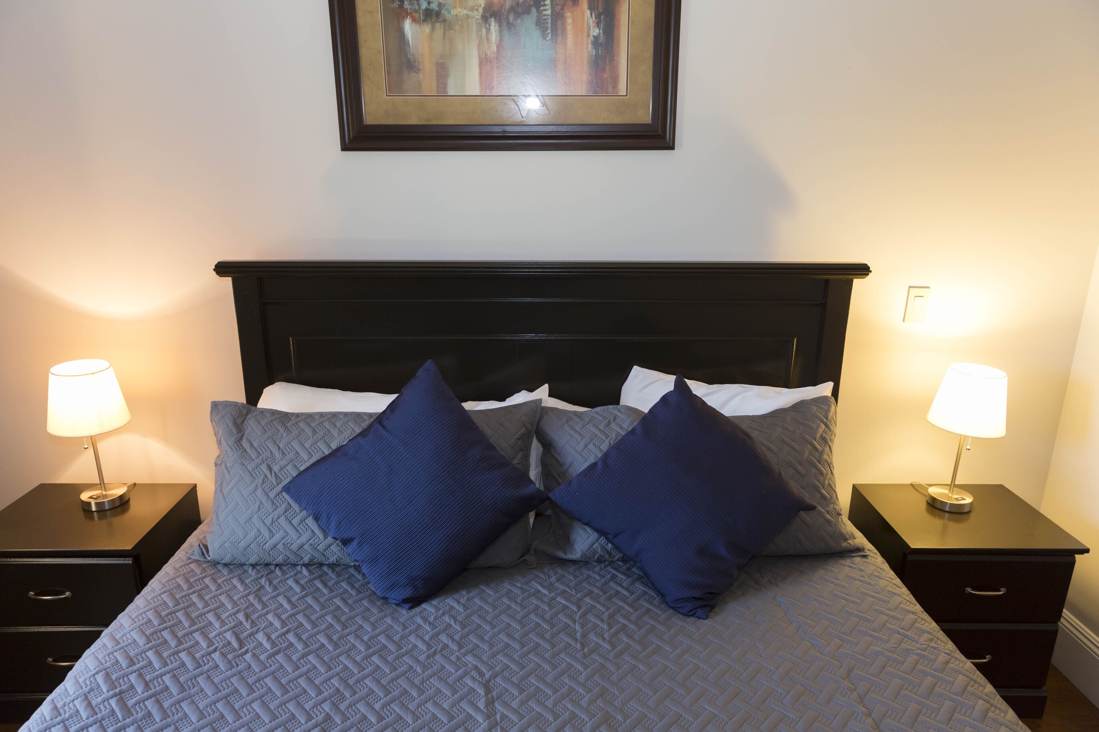 A neatly made bed with a quilted headboard, two bedside tables with lamps, and a framed artwork above the bed.