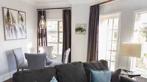 Una sala moderna con un sofá seccional gris, una mesa de centro de vidrio y un sillón estampado. Hay dos ventanas con cortinas, una lámpara colgante y obras de arte en las paredes.