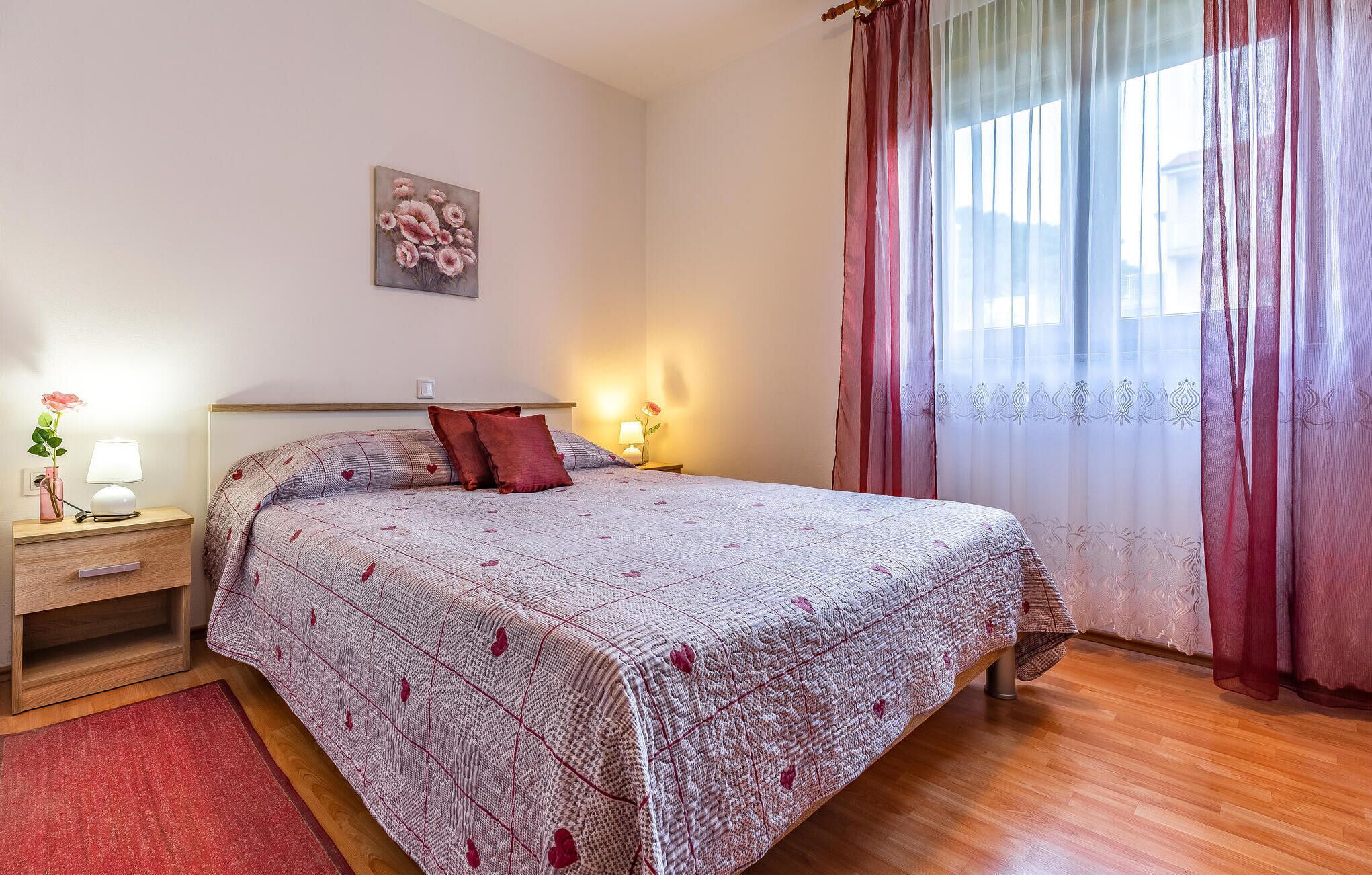 A bedroom with a bed, bedside tables, a nightstand, a lamp, a vase with a flower, and a window with curtains.