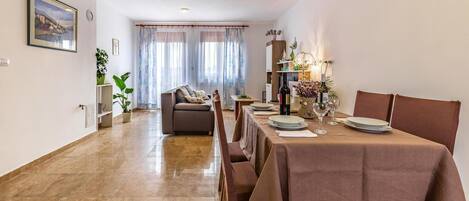 A dining area with a table set for four, a sofa, and a view of a dining room through a window with sheer curtains.