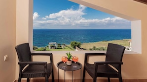 Un balcon avec deux fauteuils en osier et une petite table, offrant une vue sur la côte, la mer et un bâtiment situé au loin.