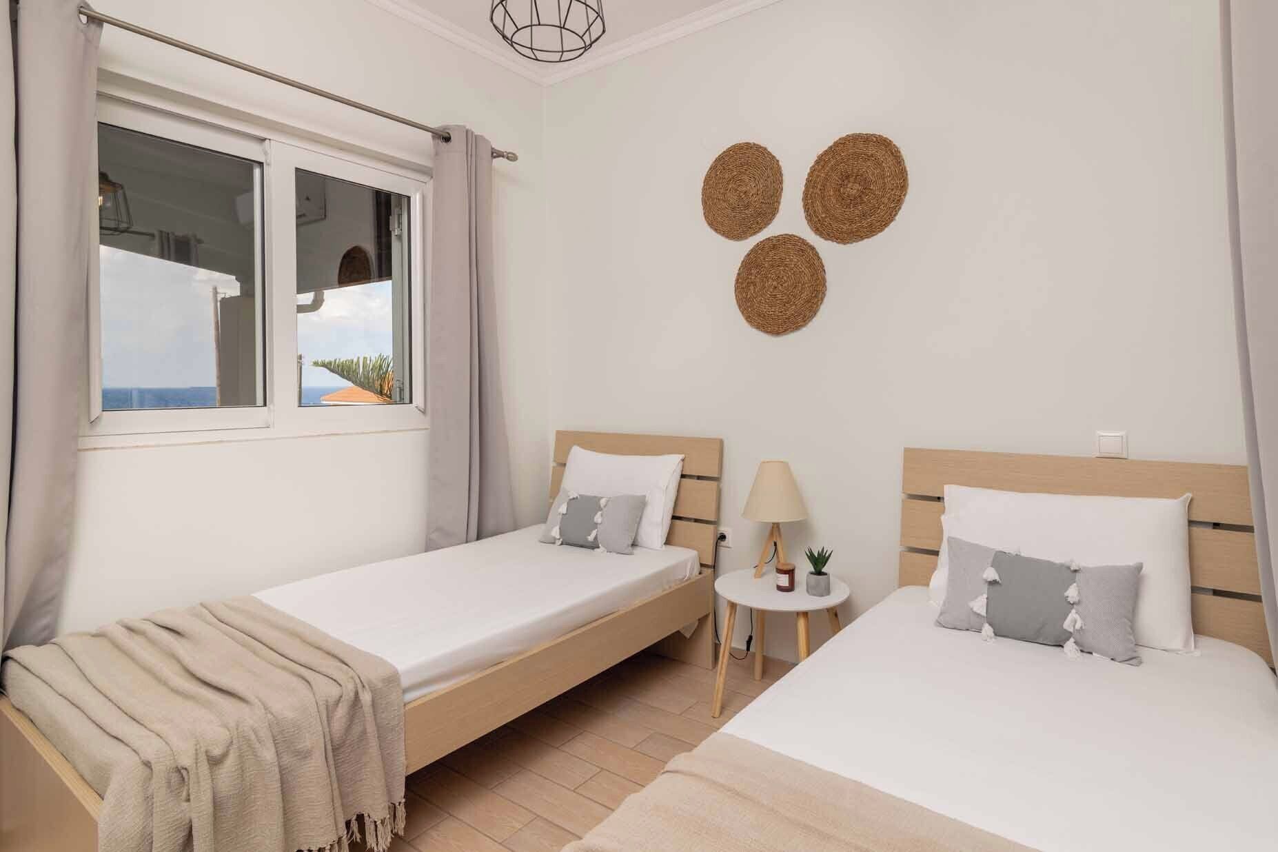 Een hotelkamer met twee bedden, een klein tafeltje met een lamp, en uitzicht op de zee door een raam.
