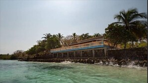 Una propiedad frente al mar con un edificio que domina el océano, palmeras y un litoral rocoso.