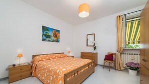 A bedroom with a bed, bedside tables, a nightstand, a mirror, a window with curtains, a potted plant, and a painting on the wall.