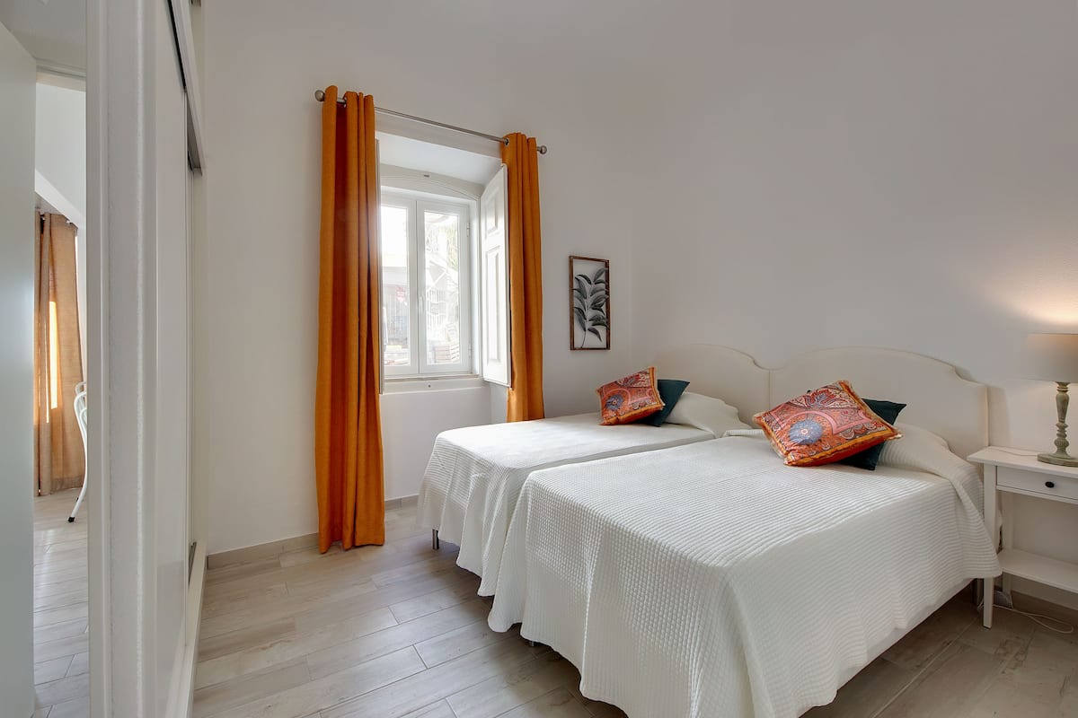 A bedroom with two beds, a window with curtains, a bedside table with a lamp, and a framed picture on the wall.