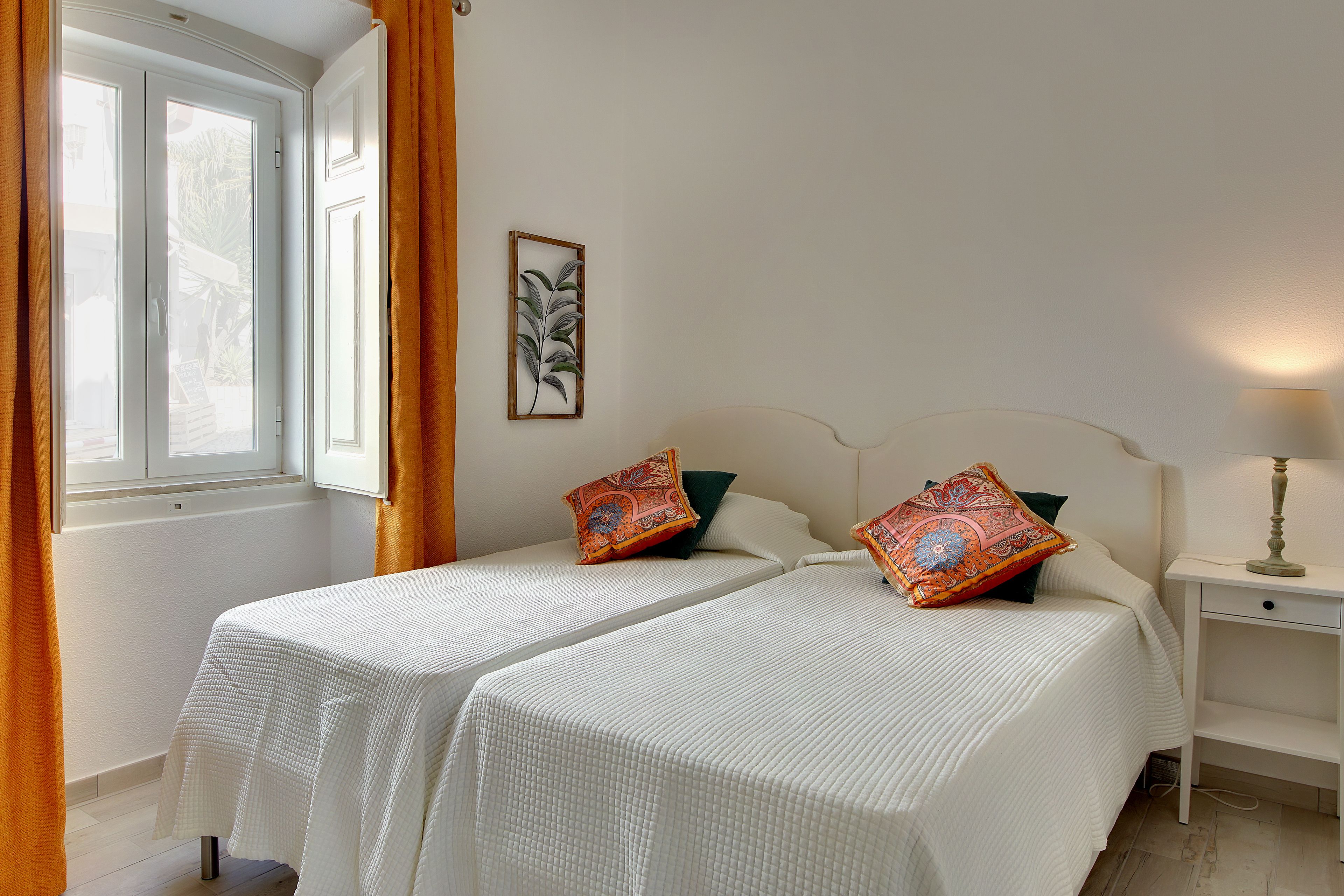 Una habitación con dos camas, una ventana con cortinas, una mesita de noche con lámpara y una planta en la pared.