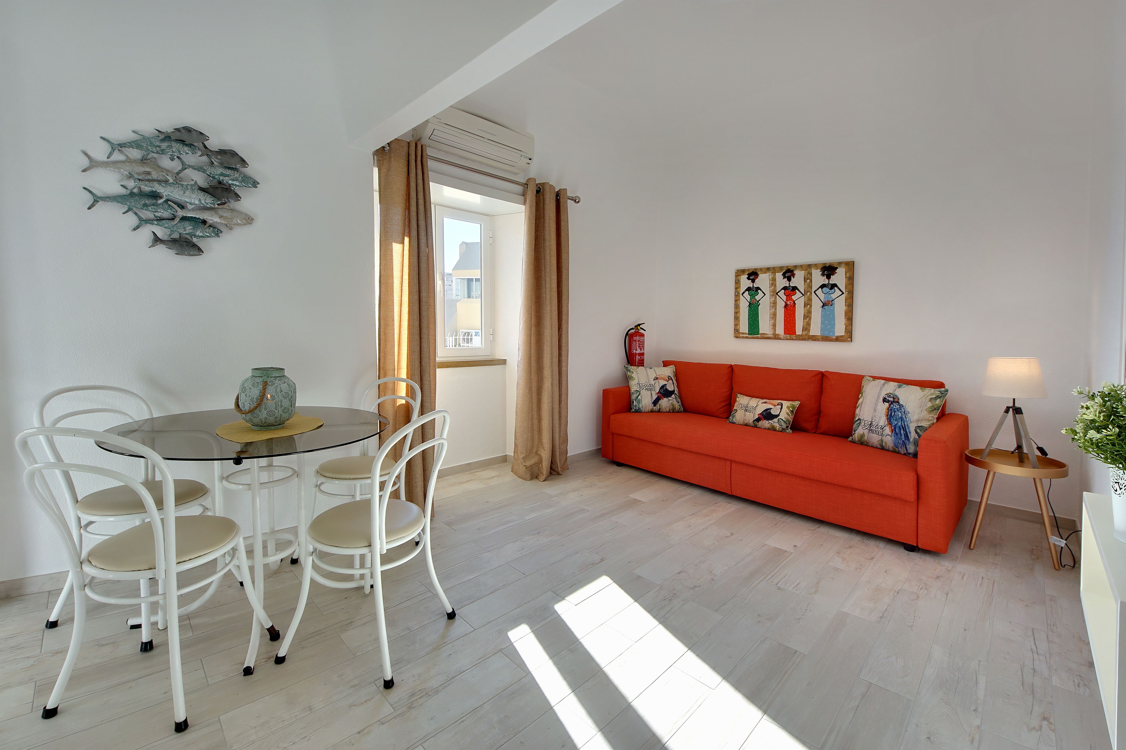 A modern living room with a round glass table, white chairs, an orange sofa, and a wall art piece.