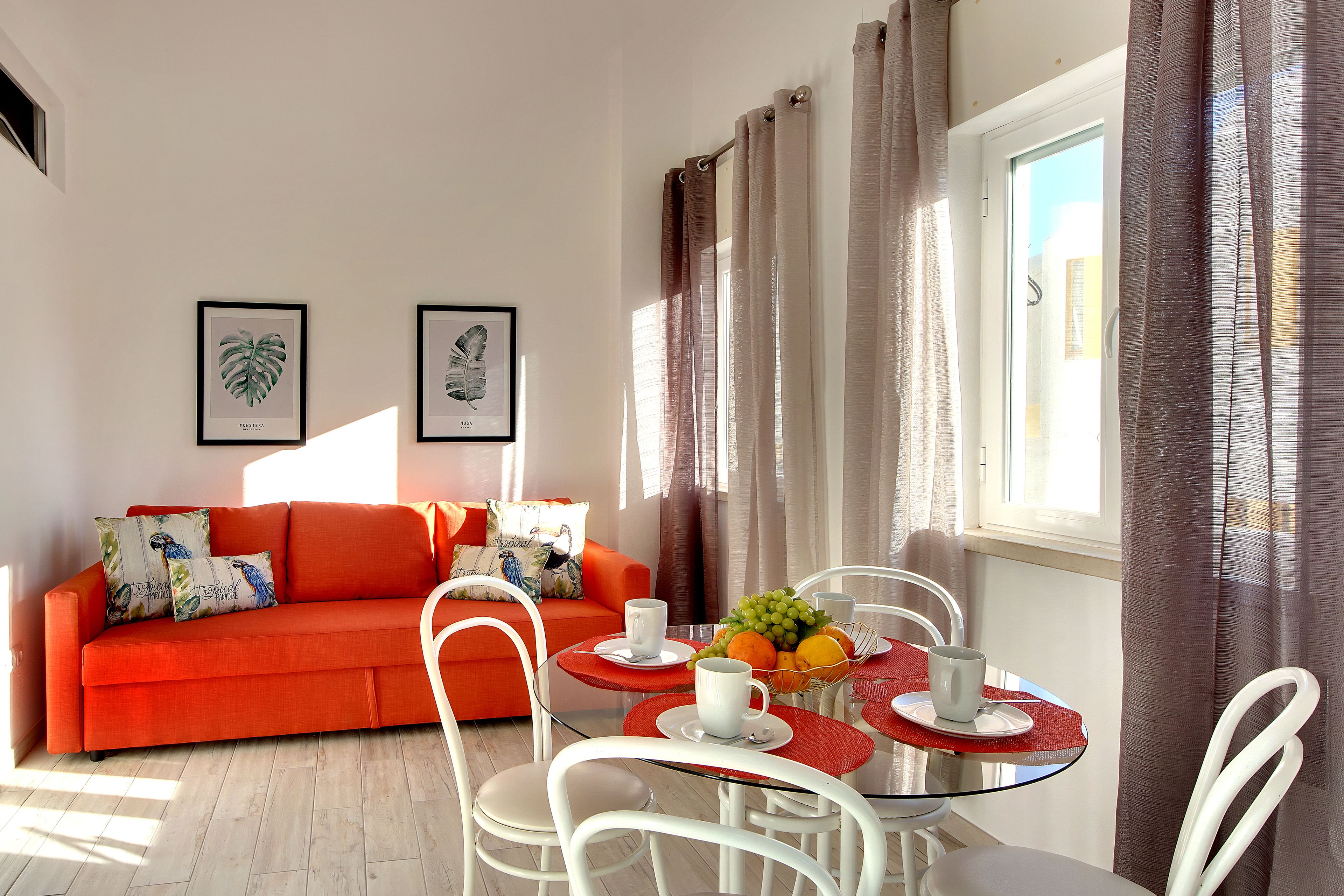 Un salón luminoso con un sofá rojo, sillas blancas, una mesa de vidrio con individuales rojos y una ventana con cortinas.