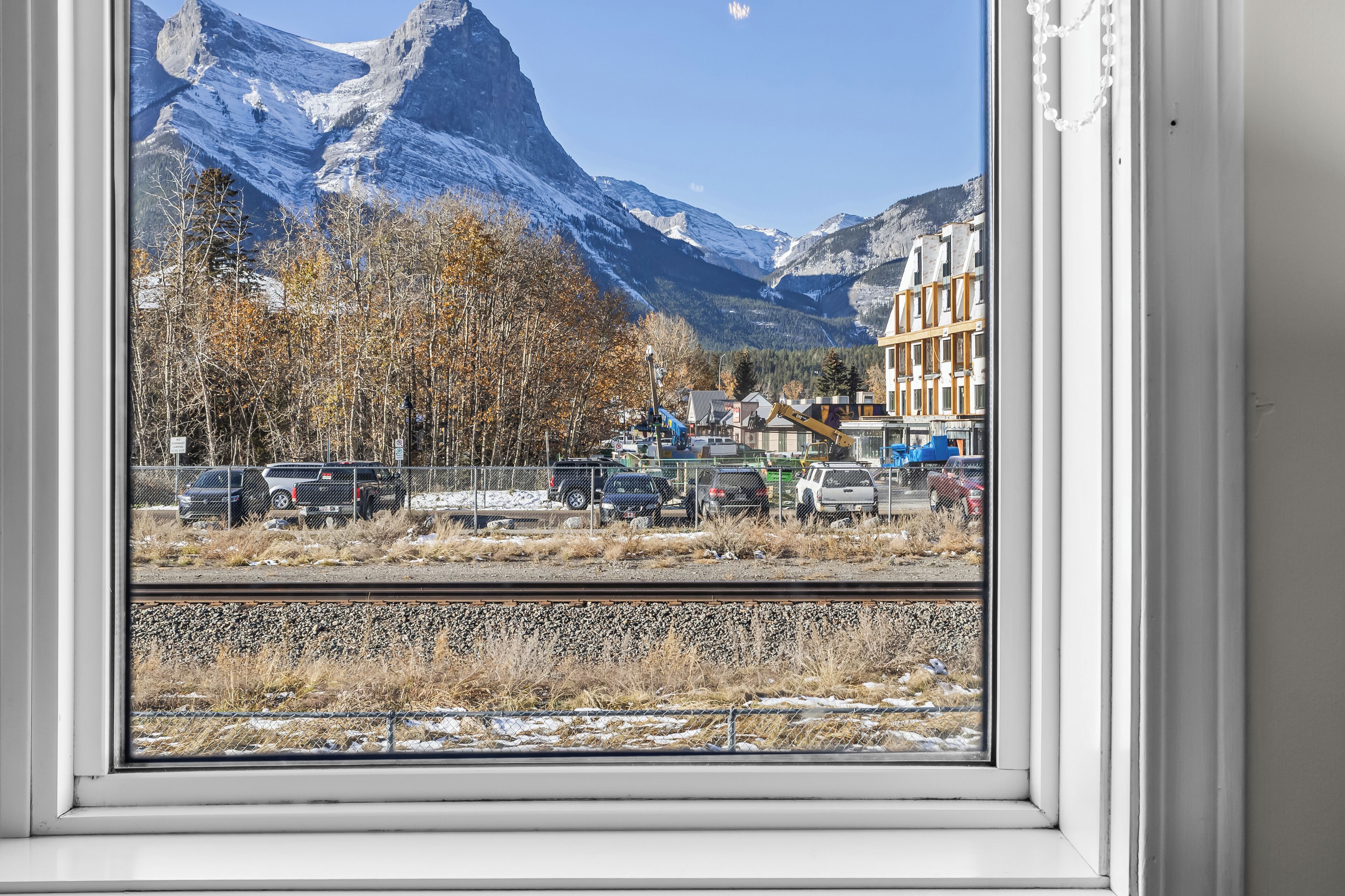 A window view of a mountainous landscape with snow-capped peaks, a parking area with several vehicles, and buildings in the foreground.