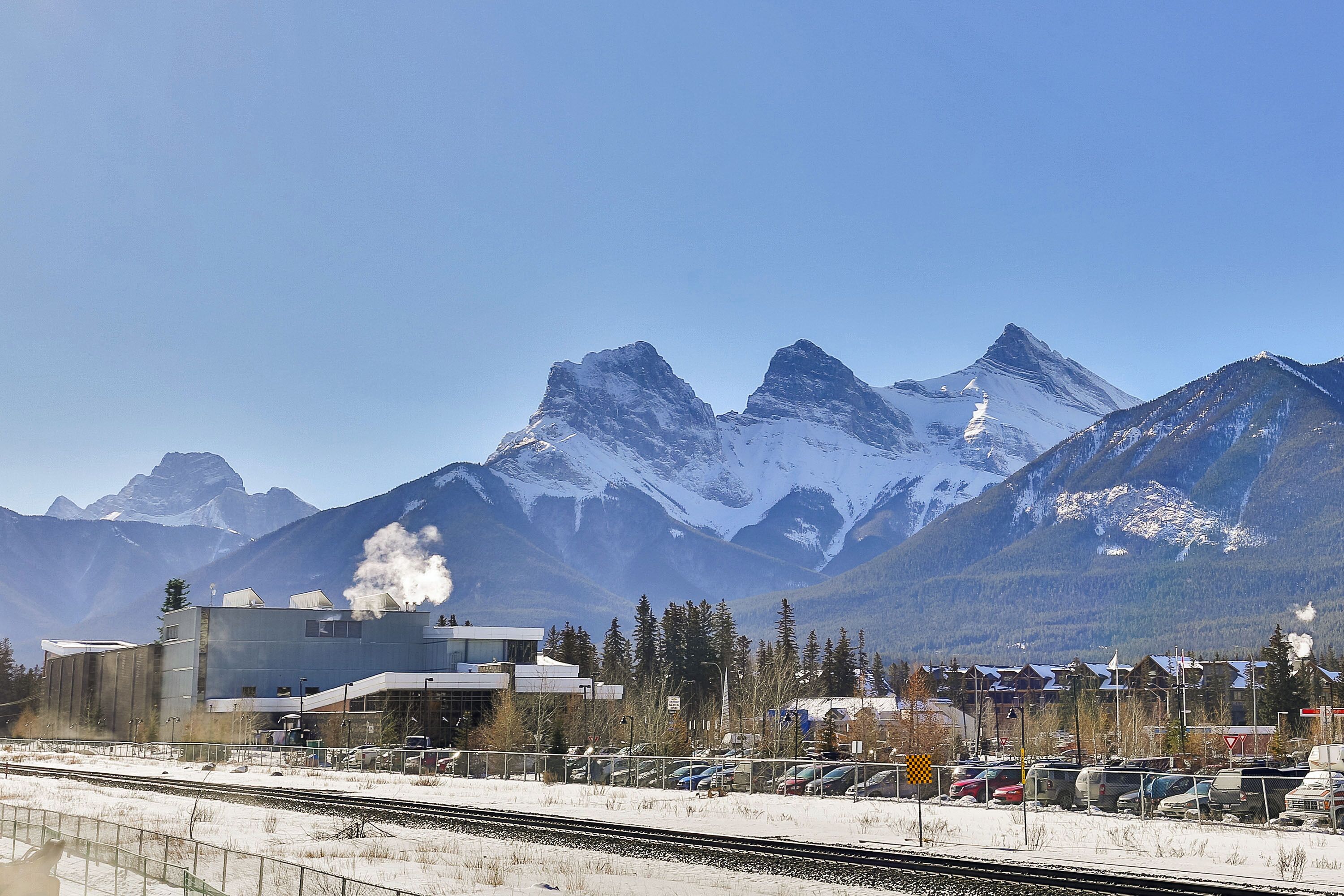 Une gare dont le bâtiment laisse échapper de la vapeur, entourée de montagnes enneigées et disposant d’un parking avec des voitures.