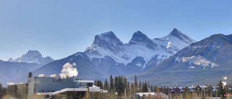 Une gare dont le bâtiment laisse échapper de la vapeur, entourée de montagnes enneigées et disposant d’un parking avec des voitures.