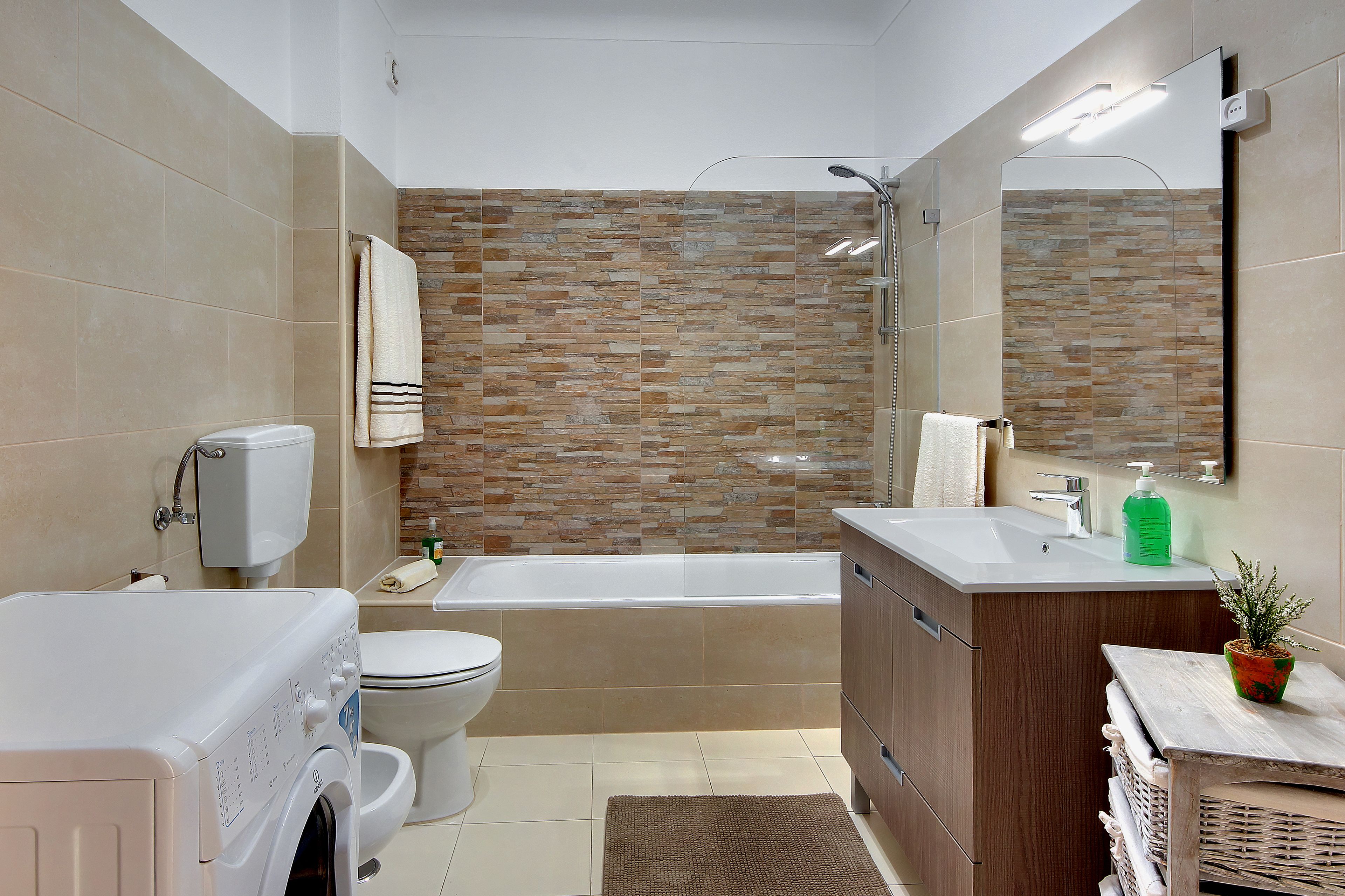 A modern bathroom with a beige tiled wall, a white bathtub, a wooden vanity, and a washing machine.