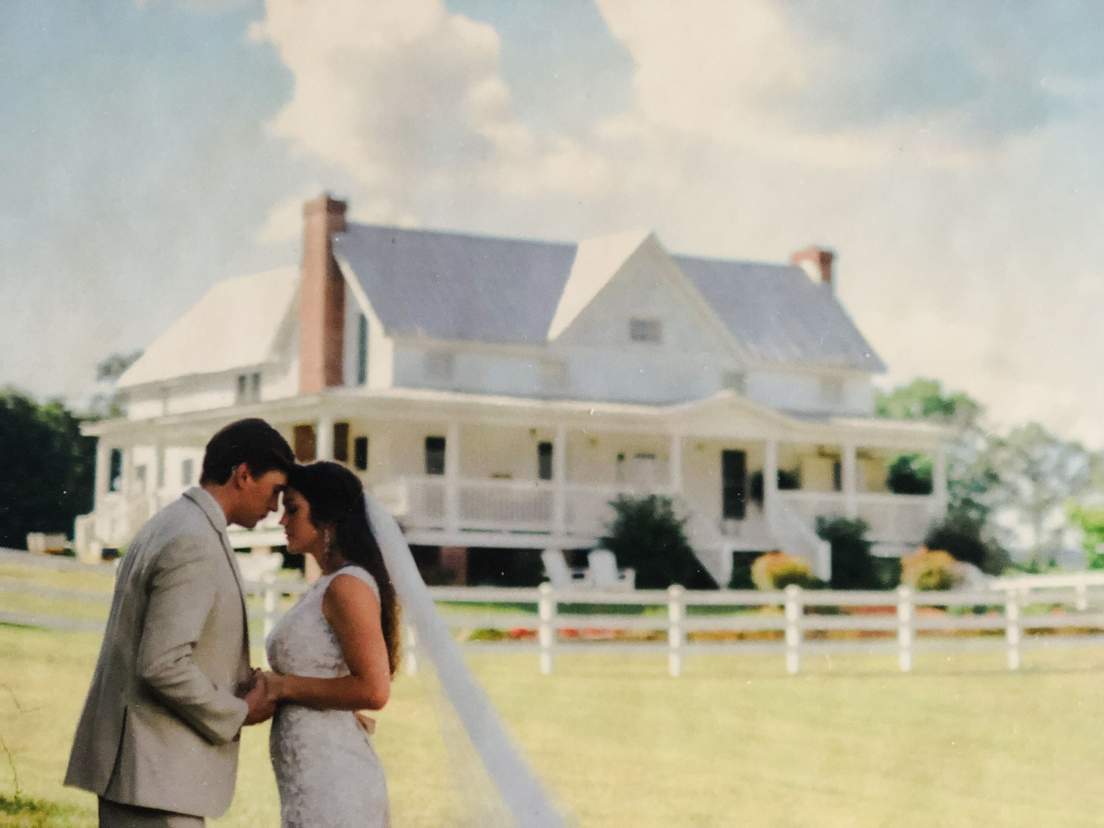 A couple embracing in front of a large, white house with a porch and a white fence.