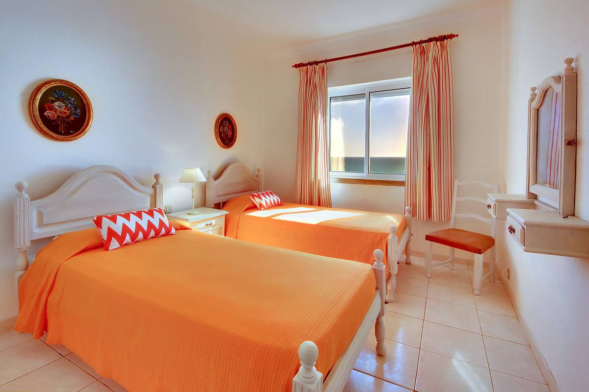 A bedroom with two beds, a bench, a nightstand, a lamp, a mirror, and a window with curtains.